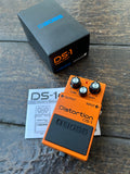 Boss DS-1 distortion pedal on a wooden surface with its packaging and manual.