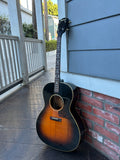 A classic acoustic guitar with a sunburst finish leans against a brick wall on a wooden porch.