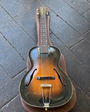 A vintage Gibson arch-top guitar with a brown sunburst finish, resting on a brown guitar case over a dark wooden plank floor.