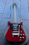 An Epiphone Wilshire red electric guitar with a glossy cherry finish, two black pick ups, nickel hardware, resting inside a black hard case on a dark gray tiled floor.