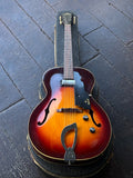 A Guild vintage arch-top  electric guitar with a sunburst finish, chrome hardware, rosewood fretboard, resting inside its black hard case on a dark wooden plank floor.