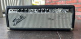 A Fender Bassman 100W guitar amplifier head, featuring a black textured cabinet, silver grille, and black knobs, photographed outdoors on a stone surface with a brick wall background.