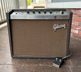 A vintage Gibson guitar amplifier model 19-RVT with a dark wood cabinet, brown grille cloth, and chrome controls, shown on a tiled floor with a brick wall in the background.
