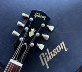 Close-up of a Gibson acoustic guitar headstock with cream-colored tuning pegs and gold Gibson branding, set against a dark blue textured surface with a larger gold Gibson logo in the background.