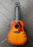 A vintage 1969 vintage Gibson J-160E acoustic guitar with a sunburst finish, rosewood bridge and fretboard, black headstock resting inside a black hard case on a dark wooden plank floor.