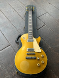 A 1973 Gibson gold top Les Paul Deluxe electric guitar with a cream pickguard and gold hardware, resting on top of a black hard case on a dark wooden floor