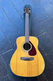 A Yamaha natural wood acoustic guitar with a dark reddish-brown pickguard and black headstock, resting on top of its black hard case against a dark blue wooden floor.