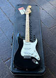 A black Fender Stratocaster electric guitar with a white pickguard and maple neck, resting inside its black hard case on a dark wooden plank floor.