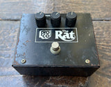A black metal guitar pedal with three black knobs, a silver footswitch, and a white 'Pro Co. The Rat' logo on the top, photographed on a rustic wooden surface.