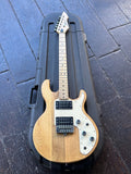 A Peavey T-25 natural wood electric guitar with a cream pickguard and black hardware, resting diagonally on top of a black hard case over dark wooden planks.