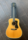 A vintage 1984 Guild acoustic guitar with a natural wood finish and black pickguard, ebony fretboard, black headstock resting vertically on a dark wooden floor next to its black hard case.