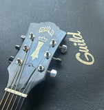 Close-up of a Guild acoustic guitar headstock with six silver tuning pegs and six strings, resting on a black textured surface with gold Guild branding.