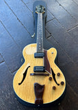 A Fender D'Aquisto double-cutaway archtop electric guitar, rosewood bridge and ebony fretboard, with a natural flamed maple top and dark wood body, displayed diagonally on a dark wooden plank floor.