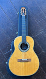 An Ovation classical acoustic guitar with a natural wood finish and a black hard case, laid flat on a dark gray tiled surface