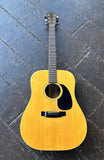 A natural wood Takamine  acoustic guitar with a black pickguard and headstock, laid flat on a dark gray stone-tiled floor.