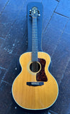A Guild acoustic guitar with a natural wood top and dark pickguard, resting inside a black hard case on a dark wooden plank floor.