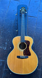 An acoustic guitar with a natural wood body and black headstock, displayed inside its black hard case on a dark blue wooden floor.