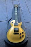 A Gibson gold top Les Paul electric guitar with black hardware and a black hard case, photographed from above on a gray tiled floor.