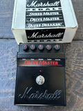 A Marshall Pedals Drive Master guitar effects pedal in black with red lettering and knobs, shown in front of its original white and black box on a dark wooden surface.