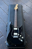 A black Stratocaster electric guitar with a maple neck and silver hardware, resting inside a black hard case on a dark wooden deck.