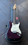 A 1999 plum purple Fender Stratocaster electric guitar resting inside its black hard case on a dark wooden floor. The guitar features a white pickguard and chrome hardware.