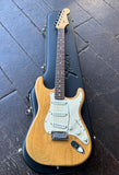A 2000 American Fender Stratocaster electric guitar in a natural wood finish with a white pick-guard, rosewood fingerboard, maple headstock resting inside a black hard case on a dark wooden floor.