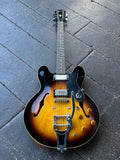 An Epiphone classic double-cutaway electric guitar with a sunburst finish with chrome hardare, bibsgy tremolo, resting on a dark gray wooden plank floor. The guitar features a black headstock, black pickguard, and chrome hardware, including a prominent tremolo bridge.