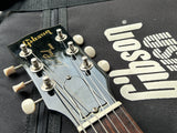 Close-up of a Gibson acoustic guitar headstock with tuning pegs and strings, resting against a black Gibson carrying bag with a large white Gibson logo.