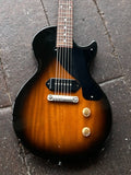A classic electric guitar with a sunburst finish, resting on a dark stone surface. The guitar features a black pickguard, black headstock, gold-toned hardware, and a brown-to-black gradient body.
