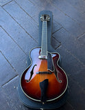 A Eastman hollow-body electric guitar with a sunburst finish, ebony fretboard resting inside a black hard case on a dark wooden plank floor.