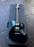 A black Fender Jaguar electric guitar with a glossy finish and silver hardware, resting inside a black hard case on a dark wooden deck.