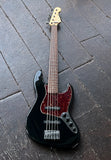 A black electric Fender five string bass guitar with a red pickguard and a light wood neck, photographed from above on a dark wooden plank floor.