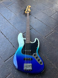 A four-string Fender Jazz bass guitar with a blue-to-white gradient finish, resting on a dark gray wooden plank floor.