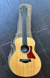 A Taylor GS Mini acoustic guitar with a natural wood finish and a red glitter pickguard, resting on its tan G.Smith branded carrying case on a dark tiled floor.