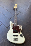 An Olympic white fender electric Jaguar bass guitar with a dark reddish-brown pickguard and gold hardware, photographed from above on a dark wooden plank floor.