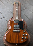 A 2014 Gibson SG in walnut finish, chrome hardware and pick ups, rosewood fret board resting inside a brown hard case on a dark wooden floor