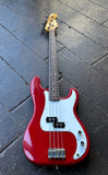 A red Fender Precision bass electric guitar with a white pickguard and gold-toned headstock, photographed from above on a dark wooden plank floor.
