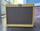 A Fender 57 vintage-style guitar amplifier with a gold textured cloth cover and a dark brown front grille, sitting on a dark tiled floor in front of a brick wall and railing.