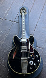 A Epiphone black electric guitar hollowbodywith chrome hardware and gold-toned trim, resting inside a black hard case on a dark wooden floor