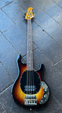A Music man Sunburst four-string bass guitar with a sunburst finish, laid flat on a dark gray stone tile surface.