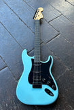 A 2025 Fender Stratocaster light blue electric guitar with a black pickguard, black pick ups, ebony fretboard and maple headstock, photographed from above on a dark wooden plank floor.