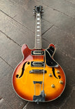 A Univox classic Coily two pick up electric guitar with a sunburst finish, resting on a dark wooden plank floor. The guitar features a warm orange-to-brown gradient body, a black pickguard, gold hardware, and a black headstock.