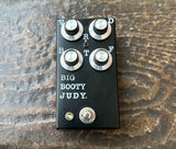A black guitar effects pedal with four silver knobs, labeled 'BIG BOOTY JUDY.' in white text, resting on a textured brown wooden surface.