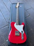 A red electric Custom S-style electric guitar with a glittery white pickguard, single pick up, rosewood fretboard, maple headstock,  resting inside a black hard case on a dark paved surface.