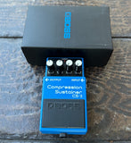 A BOSS CS-3 Compression Sustainer guitar pedal in blue and black, shown in front of its black BOSS box on a dark wooden surface.