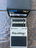 A BOSS GE-7 Equalizer guitar effects pedal shown with its black retail box on a dark textured surface. The pedal has a white body, a black control section with multiple sliders, and a black footplate with the BOSS logo.