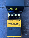 A yellow Boss OverDrive/Distortion OS-2 guitar pedal with four white knobs and a black footplate, shown in front of its black carrying case on a dark wooden surface.