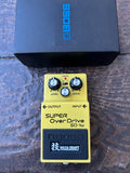 A yellow Boss Super OverDrive SD-1W guitar pedal with black knobs, shown in front of its black retail box on a dark wooden surface. The pedal features a black footplate with the Boss logo and a Waza Craft label.