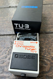 white BOSS TU-3 Chromatic Tuner for guitar on a wooden surface with packaging in the background