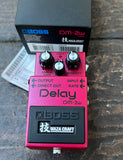 Light red Boss DM-2w Waza Craft delay pedal on a wooden surface with packaging in the background.